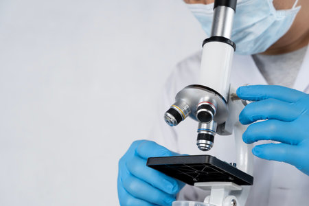 Scientist using microscope in laboratory while wearing protective gloves and mask, representing...