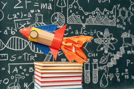 Paper rocket placed on stack of colorful books in front of chalkboard full of scientific formulas...