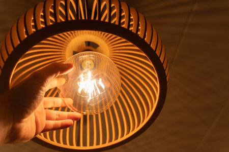 Close up of hand adjusting glowing bulb inside wooden pendant lamp, warm cozy lighting design for...
