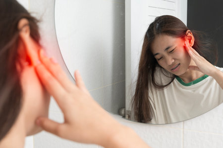 Young woman looking in bathroom mirror holding her painful ear with red highlight, showing ear...