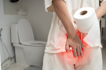 Close-up of a person with pain in the anal area holding toilet paper. Red highlight shows discomfort. Concept of hemorrhoids, irritation, anal pain, itching, or digestive health issues in the bathroomの写真素材