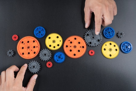Top view of colorful plastic gears and human hands assembling cogwheels on black background, symbolizing teamwork, engineering, creativity, machine parts, learning, and innovation process.の写真素材