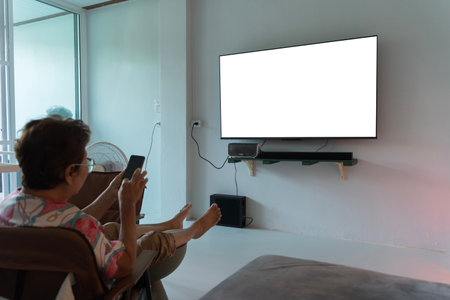 Senior woman sitting comfortably at home, holding a smartphone and facing a blank TV screen. Concept...