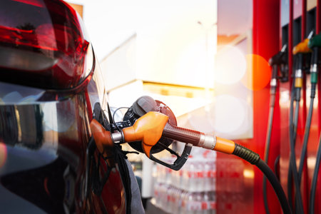 Close-up of car refueling at gas station with orange fuel nozzle, sunlight reflections, petroleum...