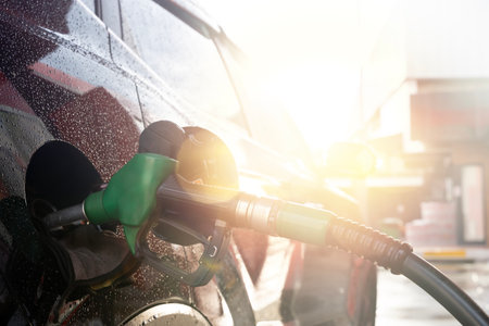 Closeup of fuel nozzle inserted in car tank at gas station with water droplets and sunlight,...