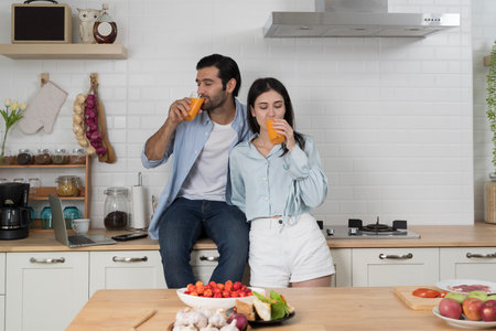 Young couple enjoying morning drink together in modern kitchen, holding glasses of fresh orange juice with laptop on counter, concept of healthy lifestyle, happiness, relationship, wellness and familyの写真素材