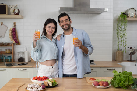 Happy young couple standing together in modern kitchen smiling and holding fresh orange juice glasses, surrounded by healthy food, concept of love, lifestyle, nutrition, wellness and family time.の写真素材