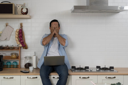 Upset young man sitting on kitchen counter covering face with hands in frustration while using laptop, concept of stress, failure, remote work problem, burnout, digital lifestyle struggle and emotion.の写真素材
