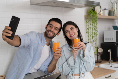 Young couple sitting together in modern kitchen, smiling and taking selfie with smartphone while holding orange juice glasses, concept of happiness, love, lifestyle, technology, health relationship.の写真素材