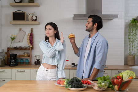Serious young woman refusing burger offered by man in modern kitchen while surrounded by fresh...