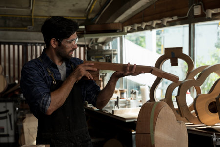 Professional carpenter inspecting handcrafted wooden guitar neck for precision and detail in workshop, wearing safety glasses and apron, representing craftsmanship, music instrument making, woodwork.の写真素材
