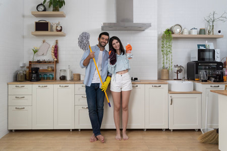 Joyful couple standing together in modern kitchen holding mop, spray bottle, and cloth while smiling, representing teamwork, happiness, fun during home cleaning, lifestyle, and domestic living conceptの写真素材