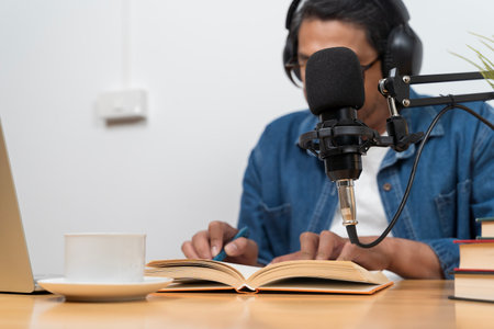 A man records audio with a microphone while reading a book and taking notes. The scene shows a podcast workspace with learning materials, emphasizing knowledge sharing, educational content creation.の写真素材