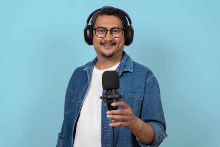 Confident man wearing headphones holding a microphone and smiling in a studio setting on a blue background, representing podcasting, broadcasting, audio recording, communication, streaming, media workの写真素材