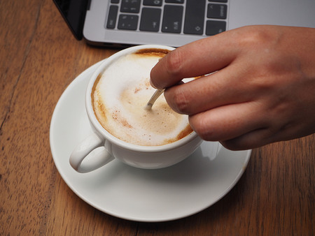 A hand stirring coffee in a white coffee cup besides a laptop on a wooden tableの写真素材