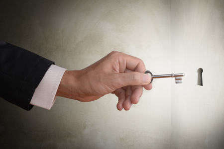 Business man hand holding key to the hole to unlock the secret, abstract business concept to solve problem, find solution, get success or find key strategy. Dark tone with concrete wall background and glow light effect.の写真素材