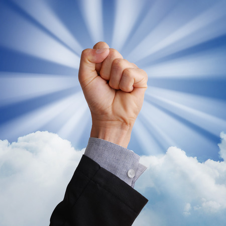 Business man hand holding fist and raising in the air in business concept in glory, happy, victory, winning, confident, or success. Blue sky, white cloud background with sun ray effect. の写真素材