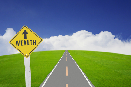 Traffic plate with text WEALTH by long road on green grass field and blue sky, white cloud background. Business concept in wealth is along way to go ahead.の写真素材