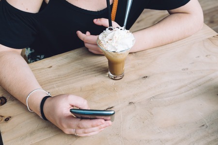 Girl drinking ice coffe and texting on her phone sitting on wooden table with space for copyの写真素材