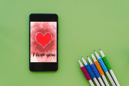Valentines day sale background with heart illustration with the words I Love You on smartphone screen mockup on green background with pencils. View from above, flat layの写真素材