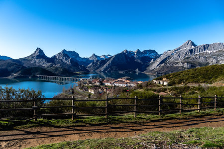 A tranquil village nestled beside a serene lake, with majestic mountains providing a stunning backdrop under a clear blue sky.の写真素材