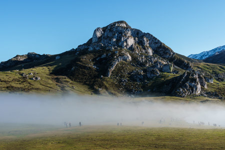 Misty morning landscape with people walking across a grassy field, a rocky mountain rising in the...