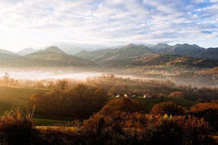 Misty morning in the valley, sun rays through the hills, autumn colors and housesの写真素材