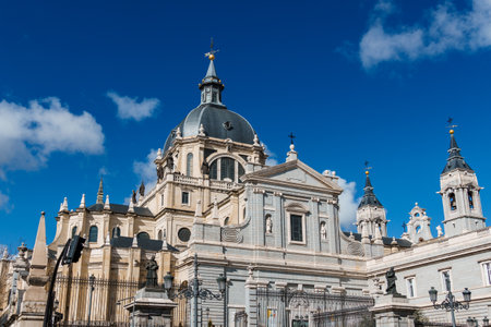 A view of the Almudena Cathedral in Madrid, Spain, featuring its detailed architecture and dome against a bright blue sky.の写真素材