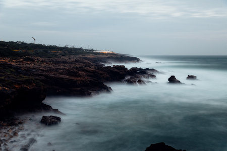 A beautiful long exposure shot shows the waves crashing against the rocky coastline of Cascais, Portugal, on a cloudy day.の写真素材