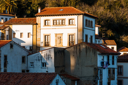 A white three-story house with red tile roof is nestled among other buildings in a hillside town, bathed in sunlight. Lastres, Asturiasの写真素材