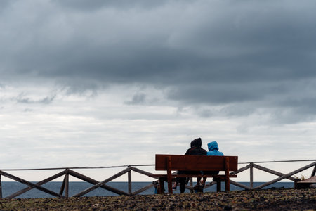 Two people sit on a bench overlooking the ocean under a cloudy sky in Sao Miguel, Azores.の写真素材