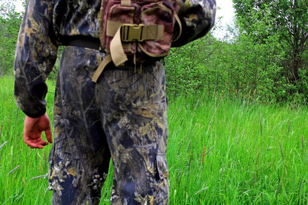 A man in camouflage with a backpack stands in a green meadow.の写真素材