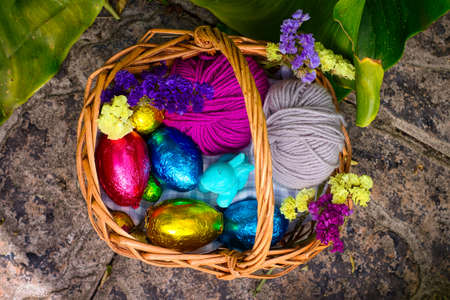A basket decorated for an egg hunt. It has eggs wrapped in color tin foil, a toy of a rabbit and some balls of yarn. It's surrounded by vegetation, clovers and yellow flowers.の写真素材