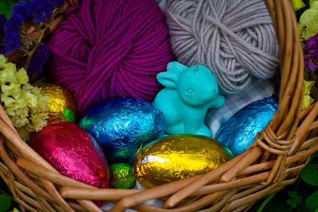 A basket decorated for an egg hunt. It has eggs wrapped in color tin foil, a toy of a rabbit and some balls of yarn. It's surrounded by vegetation, clovers and yellow flowers.の写真素材