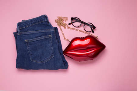 Red clutch bag shaped as woman mouth lips and folded denim jeans pants on pink background, sexy...