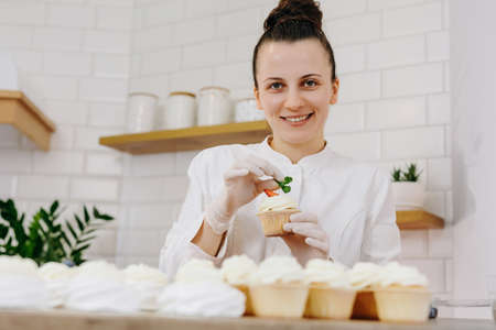 Portrait of smiling female professional decorating cupcakes with strawberry. Various delicious cakes lying on the table. Lifestyle.の写真素材
