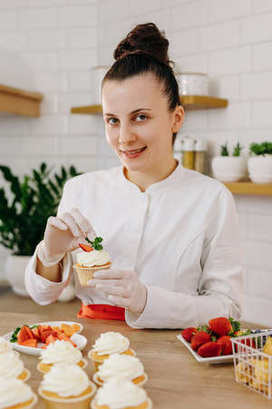 Portrait of smiling female professional decorating cupcakes with strawberry. Various delicious cakes...