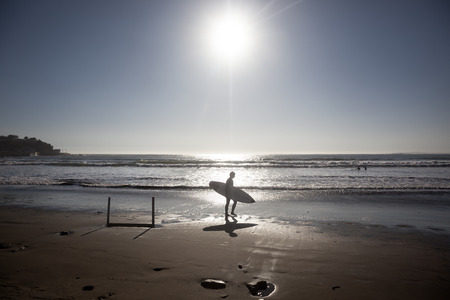 Surfer walks along the beach after a fun time in the water. Image is backlit and features the subject in silhouette.の写真素材
