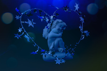 Figurine of a sleeping angel. Around the cherub is a Christmas silver decoration in the shape of a heart with stars. On the blue background are colorful blurred defocused dots soft lights  with selective focus.の写真素材