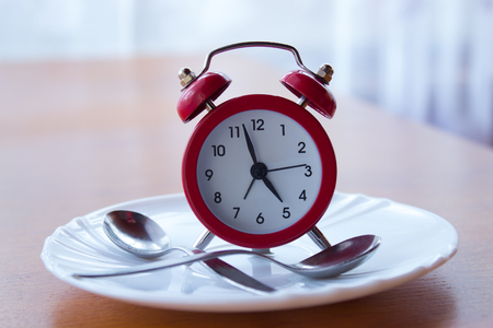 Red alarm clock on a plate with spoons stands on a bedside table. In the background  is a window with curtain. の写真素材