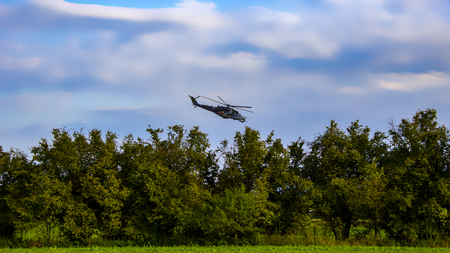 Combat helicopter on blue sky with white clouds. Trees and bush with grass.