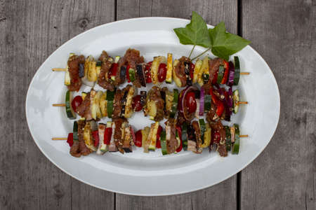 Skewer before grilling composed of bacon onions tomatoes meat pork zucchini peppers on a plate with leaves on a wooden tableの写真素材