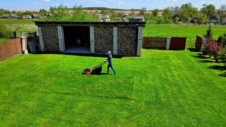 A girl in blue jeans and a sweater and a bright blue baseball cap mows grass with a red lawn mower...