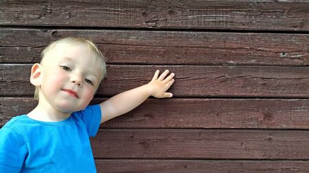 little smiling boy in a bright blue t-shirt shows something on the background of a brown wooden wall. Space for your advertisingの写真素材