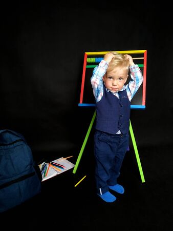 little boy in a blue suit holds his head while standing by the blackboard. back to school. Emotional boy. isolated on blackの写真素材