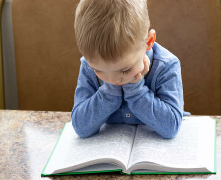 close-up of a pensive blond-haired toddler leaning over a book holding his head