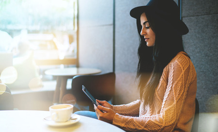 beautiful young girl watching video on digital tablet while sitting in cafe. a student in ordinary clothes and in a hat fills an online test. Coffee break. Bokeh light. Film effects.の写真素材