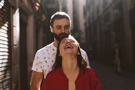 Attractive woman is laughing when her beloved man cuddles her while standing on a blurred urban background. Couple of young people in love is walking outdoors on a sunny summer day.の写真素材
