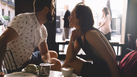 Couple of young beautiful people is sitting in a modern coffee shop on a summer day. A romantic dating of a handsome bearded guy and a charming girl in a cafe. Portrait of a man and a woman.の写真素材