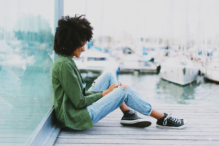 Smiling attractive freelancer female with afro hairstyle wearing jeans overalls is surfing the web on a display of a mobile phone while sitting on a bench at the city berth area on a morningの写真素材